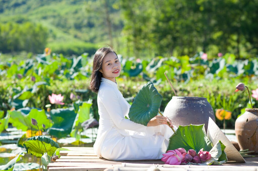Asian woman in traditional dress sitting by a lotus pond, smiling and holding green leaves.